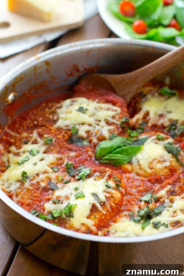 30-Minute One-Pan Chicken Parmesan 3 Close-up of crispy breaded chicken breast simmering in chunky tomato sauce, topped with melted mozzarella and fresh green basil, in a dark skillet.