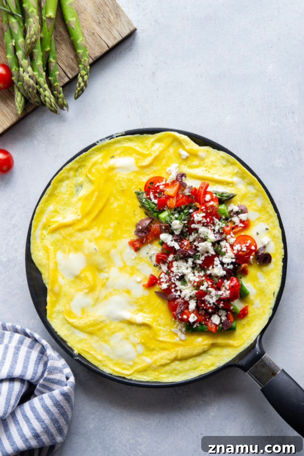 Mediterranean Asparagus & Feta Omelet 4 A close-up of a skillet with a partially folded Greek omelet, showing the rich egg texture and the vibrant fillings of asparagus, tomatoes, and feta before it's fully cooked.