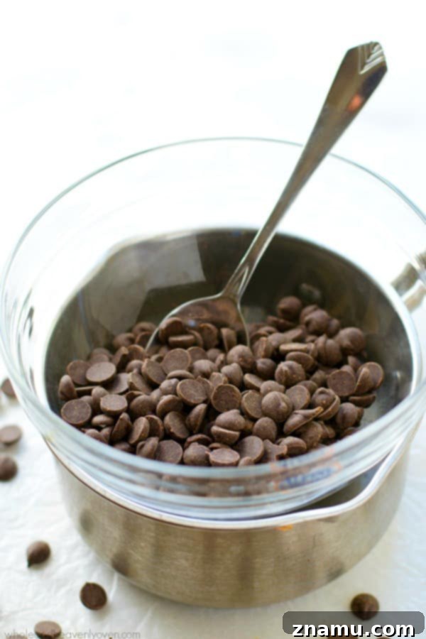 Mastering the Art of Chocolate Melting 5 A hand stirring melting chocolate chips in a glass bowl perched over a saucepan with simmering water.