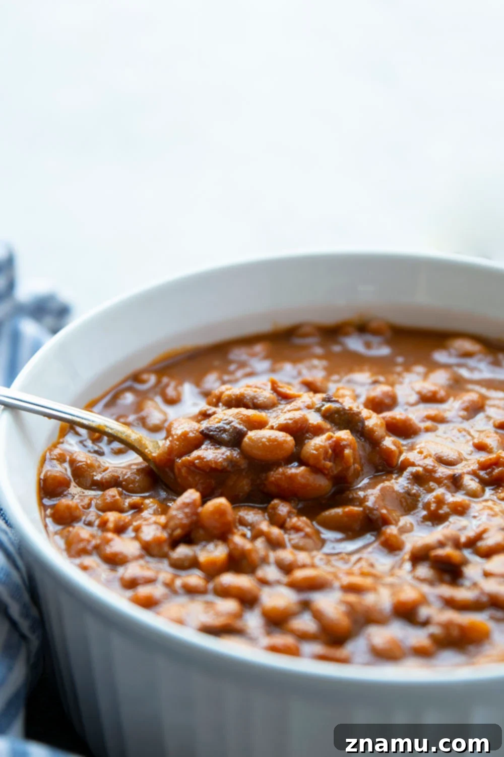 A large serving spoon scooping slow cooker baked beans from a white bowl, showing their rich, thick sauce.