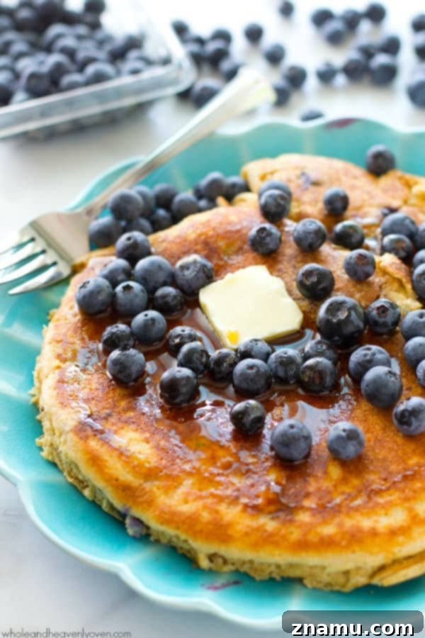 Colossal Buttermilk Blueberry Pancake 2 A towering, golden-brown XXL blueberry buttermilk pancake drizzled with maple syrup and topped with fresh blueberries.