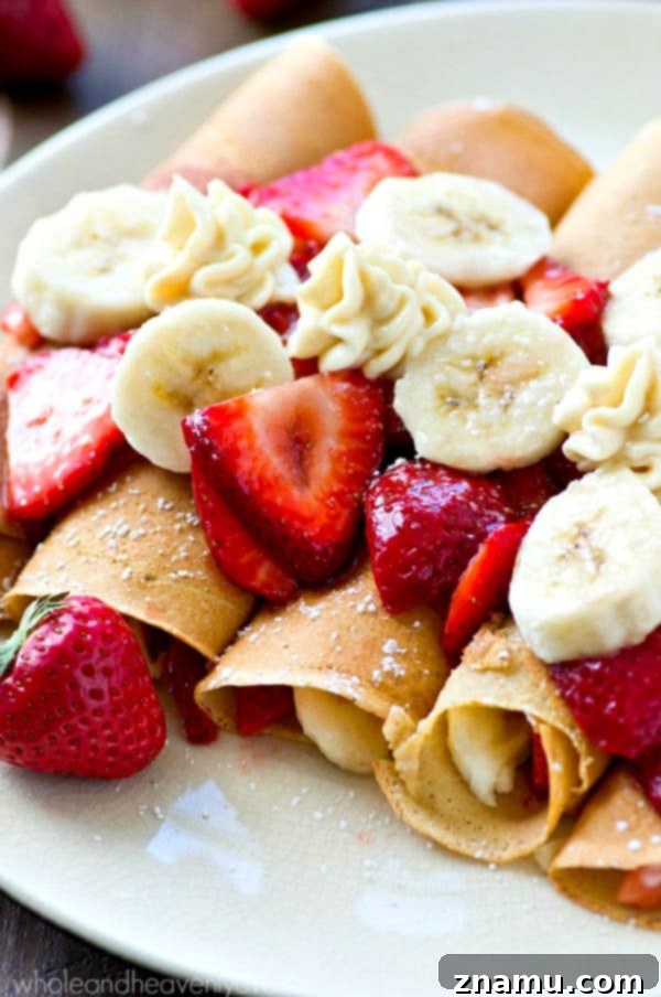 Assembled Strawberry Banana Cheesecake Crepes, generously drizzled with strawberry sauce and bananas, finished with a delicate dusting of powdered sugar.