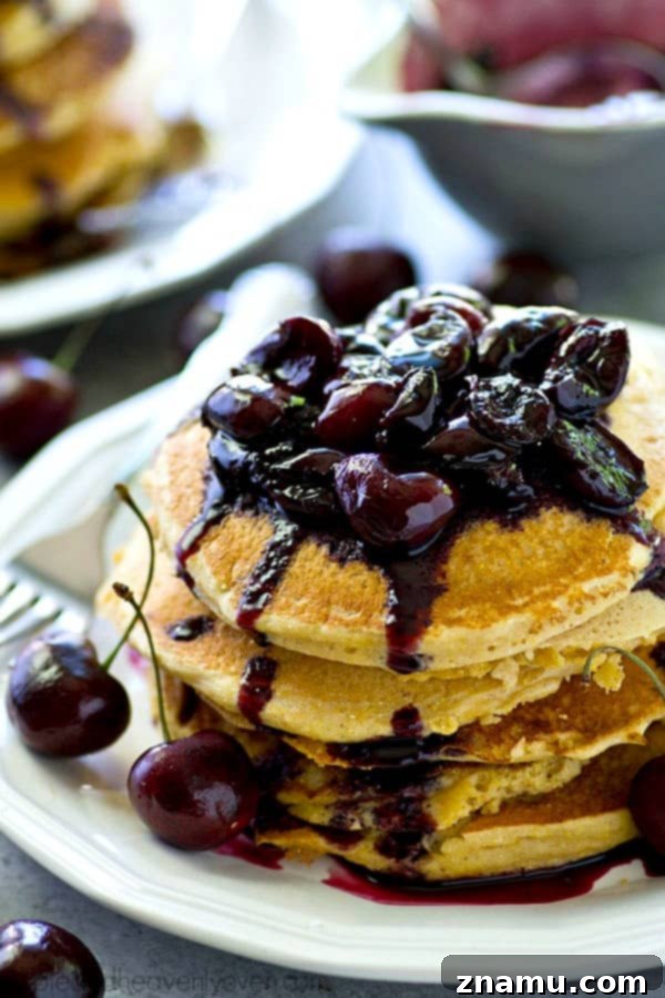 Fluffy Southern-style citrus cornmeal pancakes stacked high, generously topped with a rich, vibrant fresh cherry compote. The perfect summer breakfast.