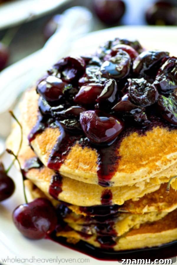 Close-up of a stack of golden citrus cornmeal pancakes with melted butter, surrounded by fresh cherries and a bowl of homemade compote.