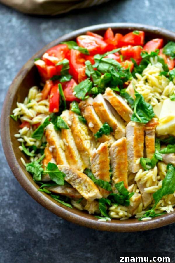 Vibrant Caprese Orzo with Grilled Chicken 3 Close-up of the Grilled Chicken Caprese Orzo Salad, showing the glistening orzo pasta coated in dressing, interspersed with succulent pieces of grilled chicken, vibrant cherry tomatoes, and soft mozzarella pearls. Fresh basil leaves add a pop of color, highlighting the fresh ingredients.