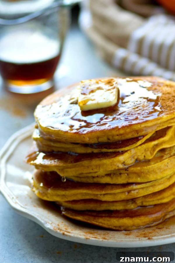 A stack of fluffy pumpkin chai spice pancakes generously drizzled with maple syrup and topped with a pat of butter.
