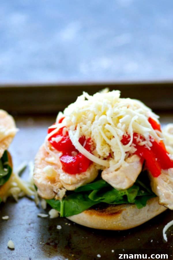 A top-down view of two freshly assembled roasted red pepper pesto chicken bagel sandwich halves, ready for broiling. Each half showcases an abundance of pesto, spinach, chicken, red peppers, and mozzarella, promising a rich and satisfying meal.