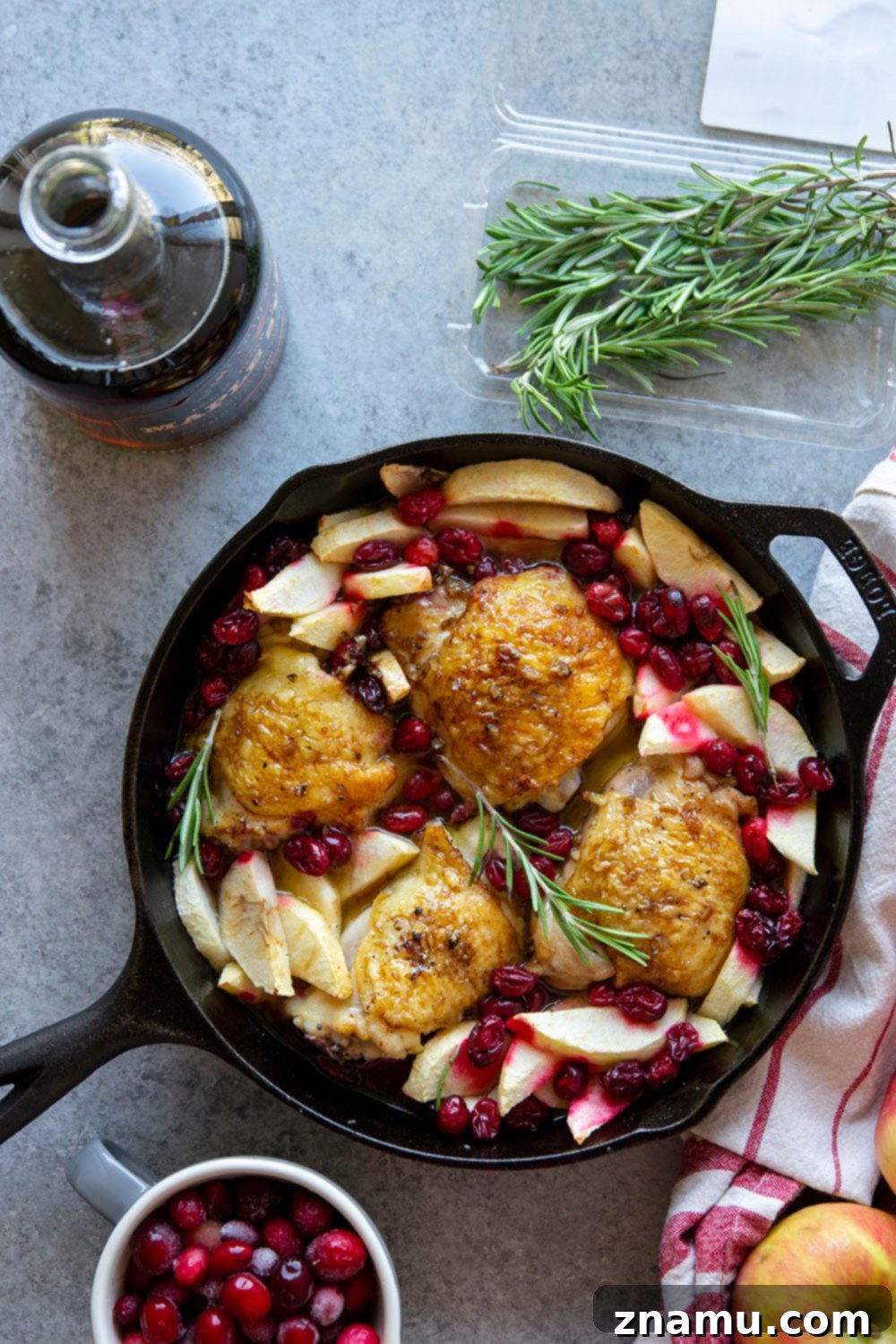 A whole maple glazed roast chicken surrounded by cranberries and apples in a skillet, ready for a festive meal.