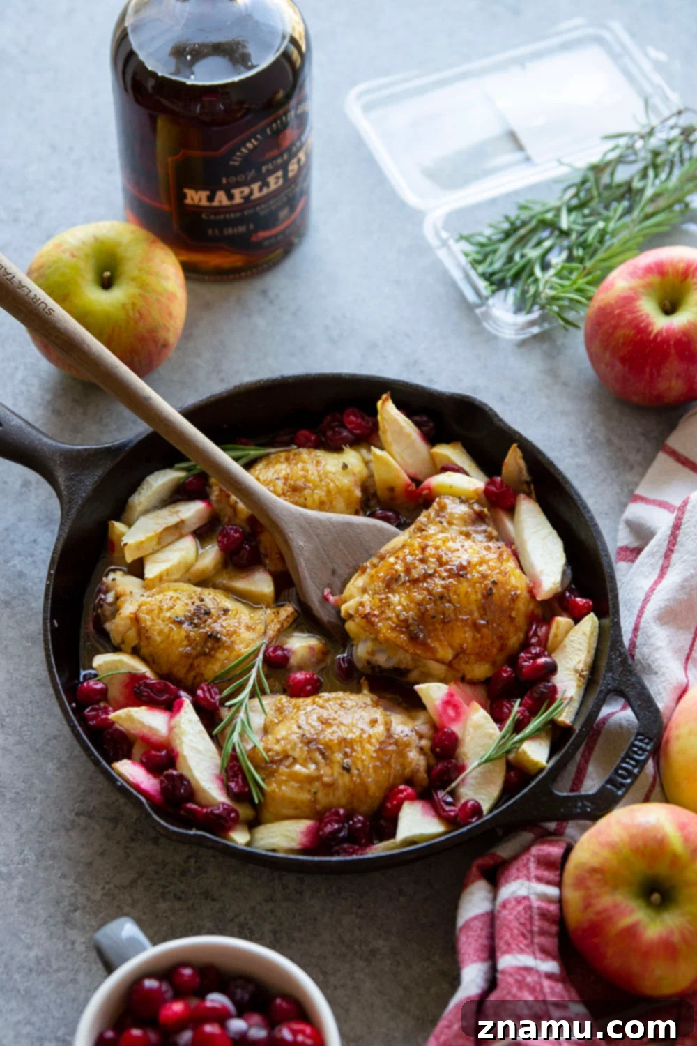 Close-up of golden-brown maple glazed chicken thighs with cranberries and apple slices in a cast iron skillet.
