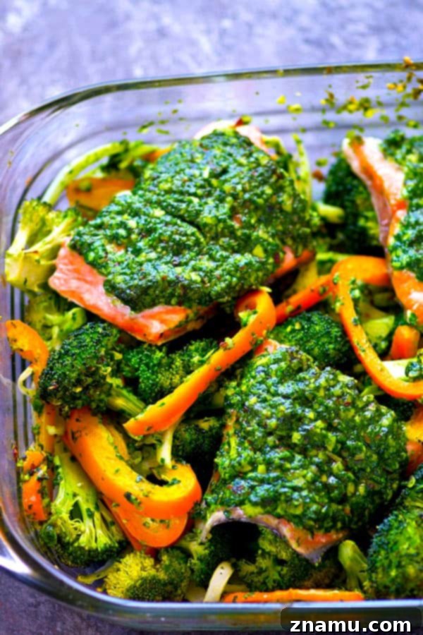 A baking sheet filled with raw salmon fillets, broccoli florets, and sliced sweet peppers, all generously coated in green pesto
