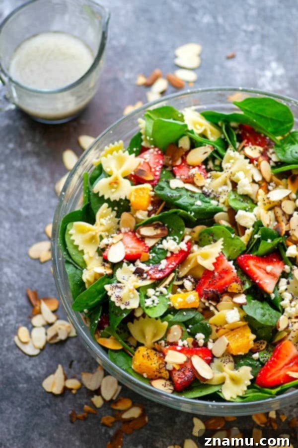 A beautifully arranged Strawberry Poppyseed Spinach Pasta Salad, featuring bowtie pasta, fresh green spinach, bright red strawberry slices, and white feta cheese, all glistening with a poppyseed dressing. The salad is presented in a rustic white bowl, ready to be served as a refreshing meal.