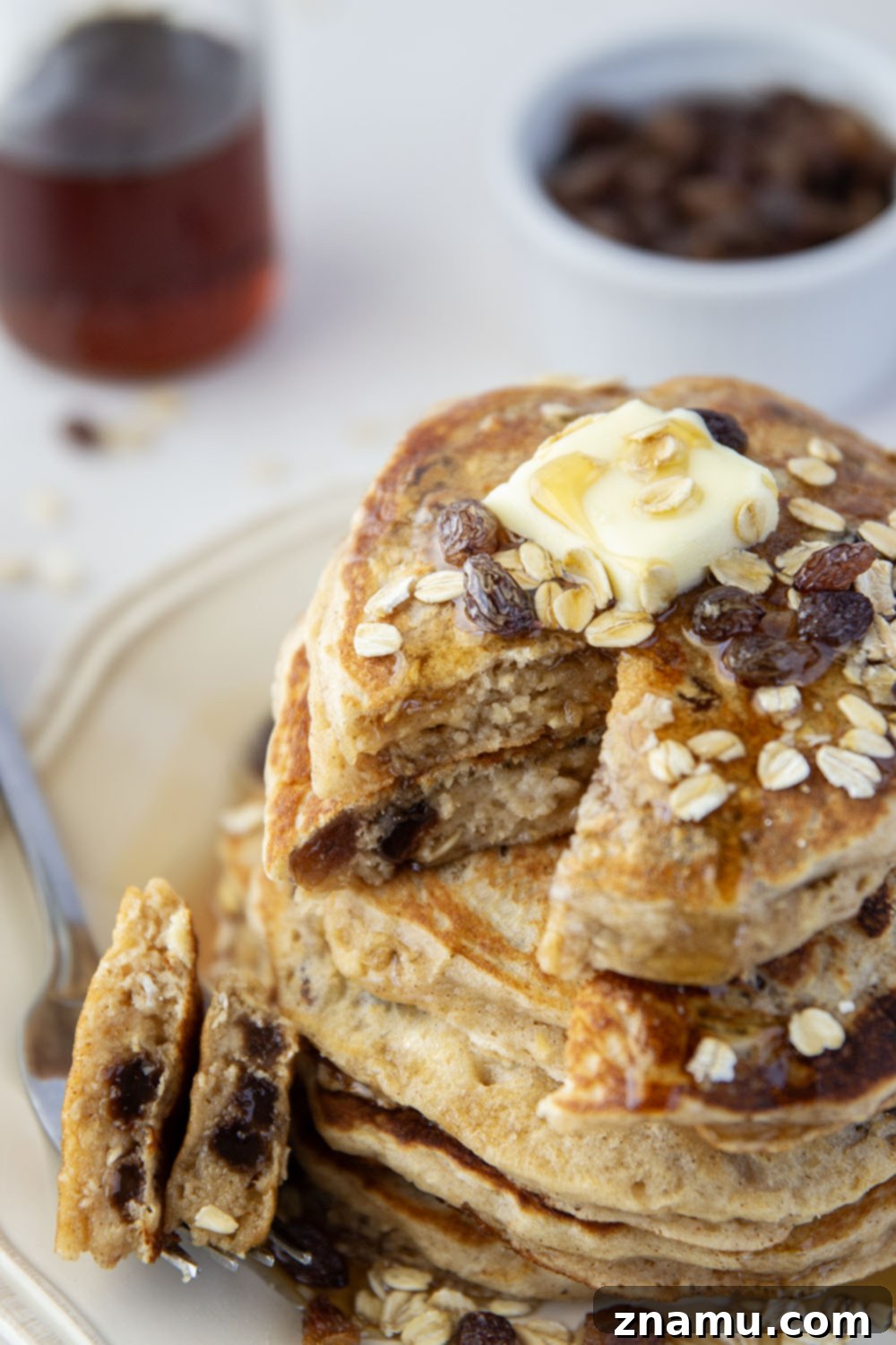 Wholesome Oat and Raisin Pancakes 6 A stack of fluffy oatmeal raisin pancakes drizzled generously with maple syrup, with a sprinkle of fresh raisins on top.