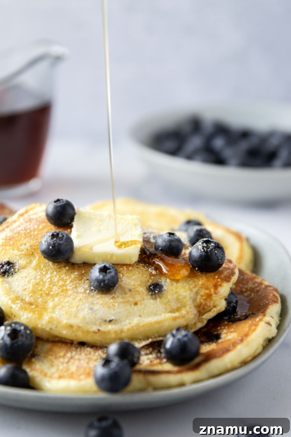 Golden Corn & Berry Flapjacks 6 A beautifully presented plate of blueberry cornmeal pancakes, garnished with fresh blueberries, a dollop of butter, and a generous pour of maple syrup.