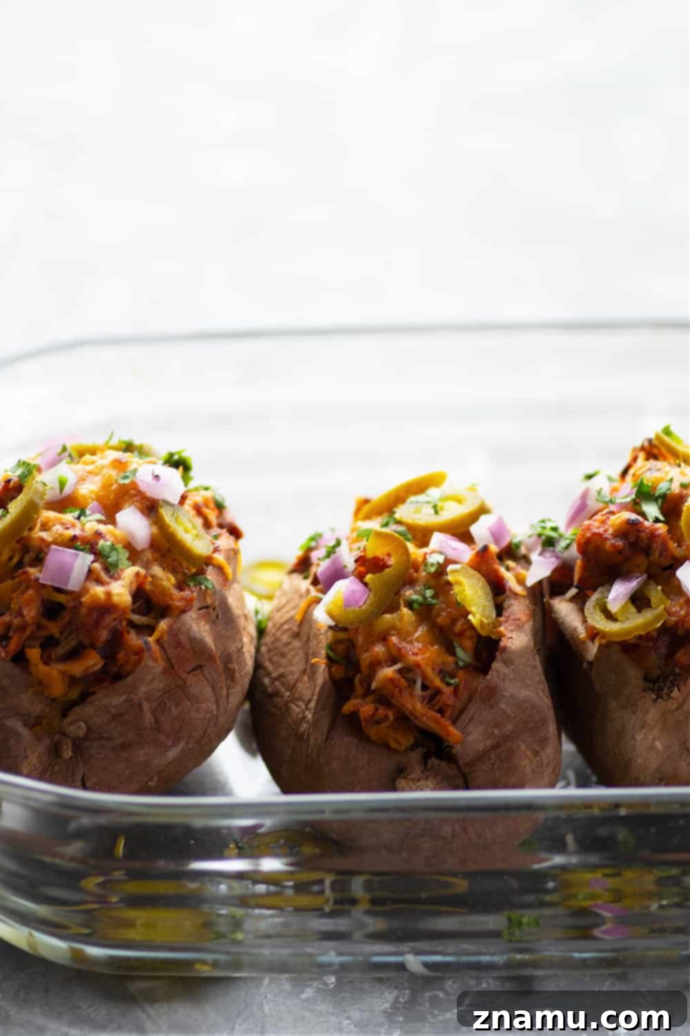 Smoky BBQ Chicken Loaded Sweet Potatoes 3 Two halves of BBQ Chicken Stuffed Sweet Potatoes on a serving plate, generously topped with fresh ingredients like red onion, cilantro, and pickled jalapenos, illustrating a complete and delicious meal.