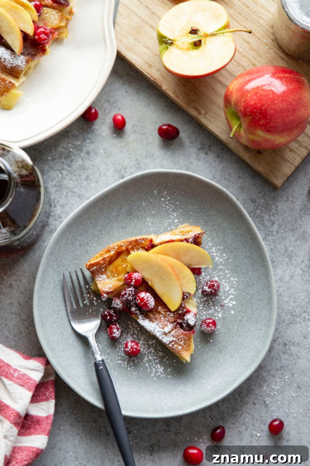 Puffed Cranberry Apple Dutch Baby 6 A slice of Cranberry Apple Dutch Baby on a white plate, showcasing the fluffy texture and colorful fruit, garnished with powdered sugar and maple syrup.