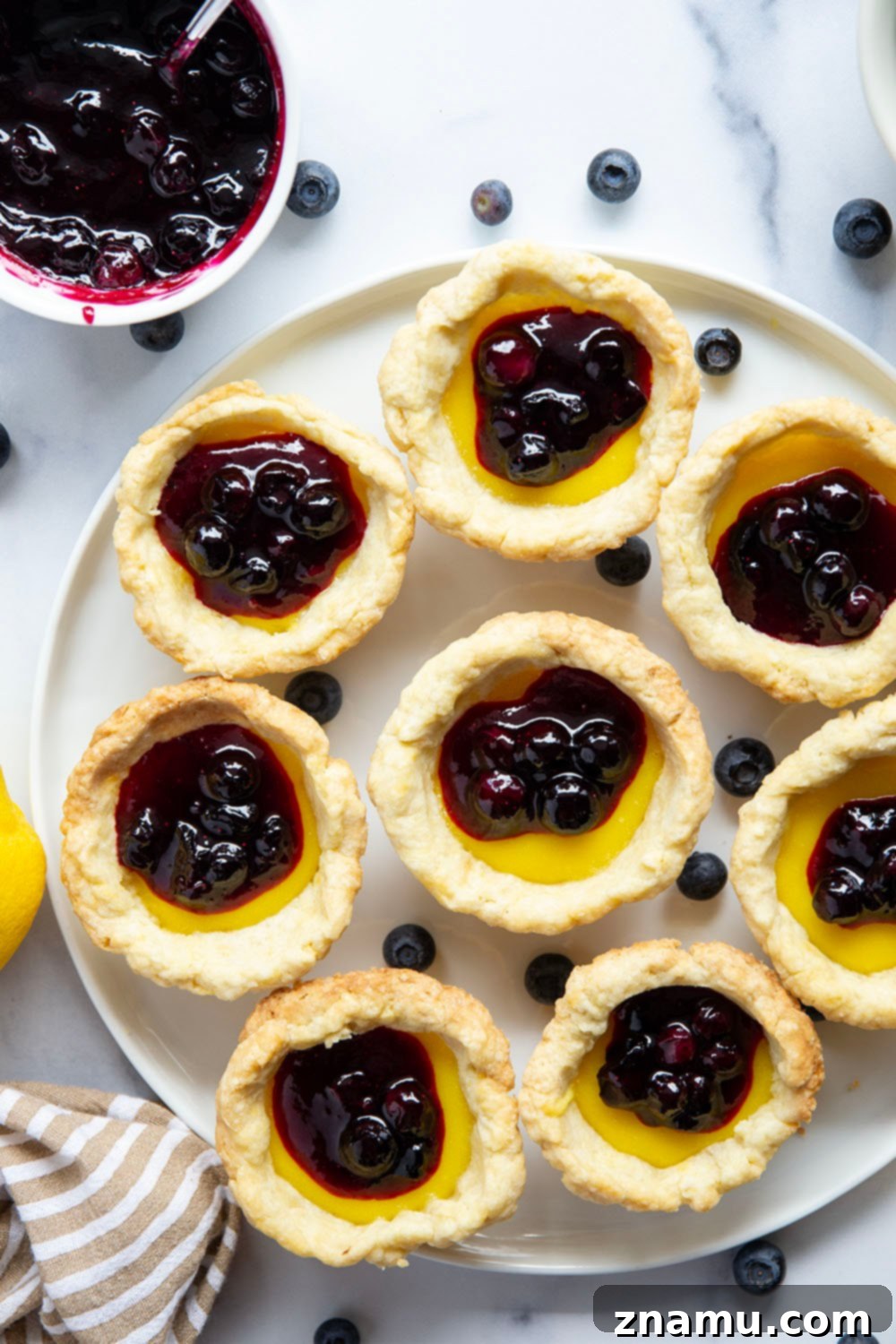 Sunshine Lemon & Blueberry Tartlets 2 Close-up of three mini lemon tarts with golden crusts, bright yellow lemon filling, and topped with vibrant blue blueberry sauce.