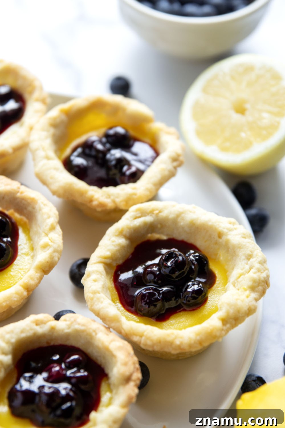 Sunshine Lemon & Blueberry Tartlets 3 Overhead view of several mini lemon tarts arranged artfully, showcasing their golden crusts and a generous dollop of blueberry sauce on each.