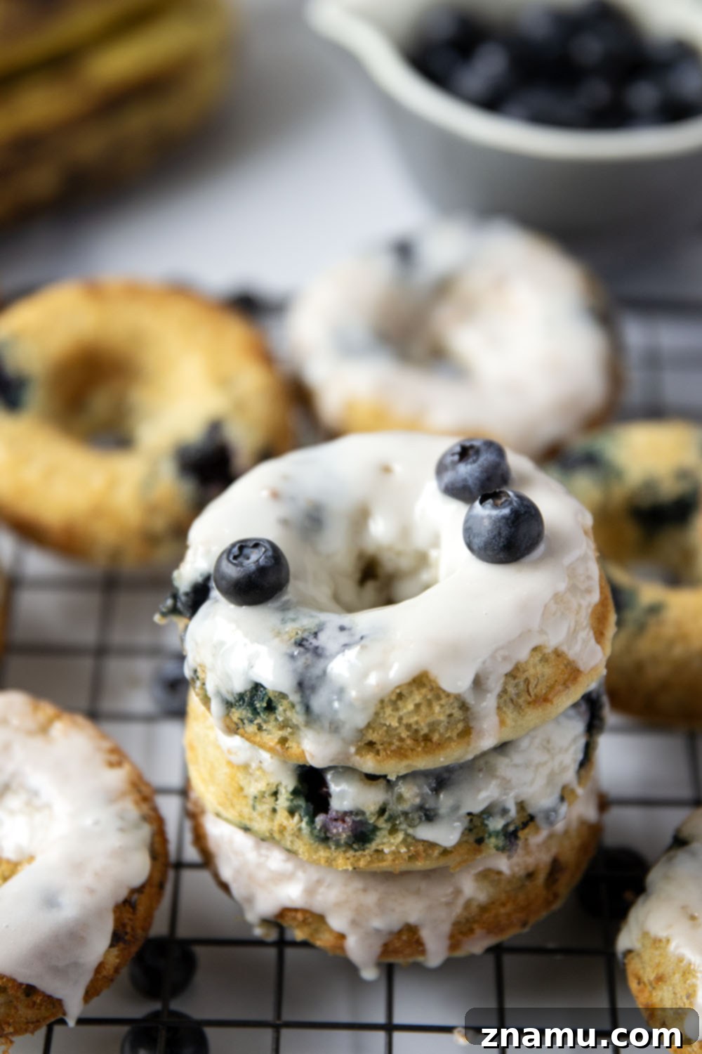 Blueberry Banana Vanilla Donuts 6 Three blueberry banana donuts, perfectly glazed with vanilla, stacked gracefully on a wire cooling rack, ready for a delicious bite.