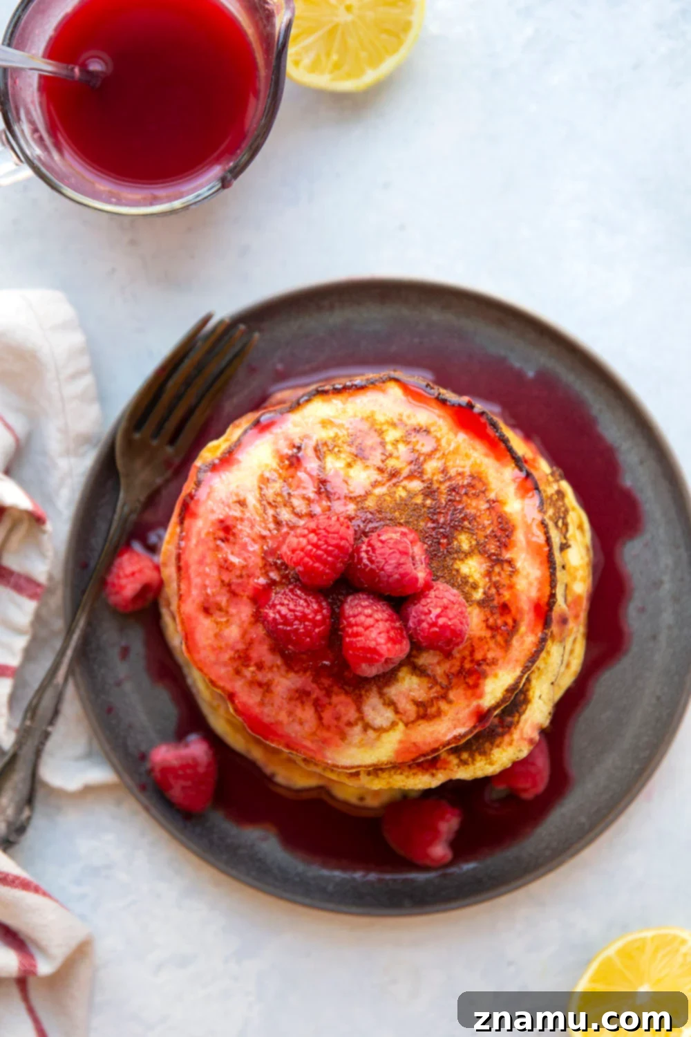 Fluffy Lemon Cottage Cheese Pancakes with Raspberry Drizzle 3 Close-up shot of a stack of fluffy lemon cottage cheese pancakes on a white plate, with a spoon gently drizzling rich, red raspberry syrup over the top. Fresh raspberries are scattered around the base, highlighting the natural ingredients. The pancakes appear thick and perfectly golden, promising a delicious and airy texture. This naturally gluten-free breakfast is made with simple, wholesome ingredients.