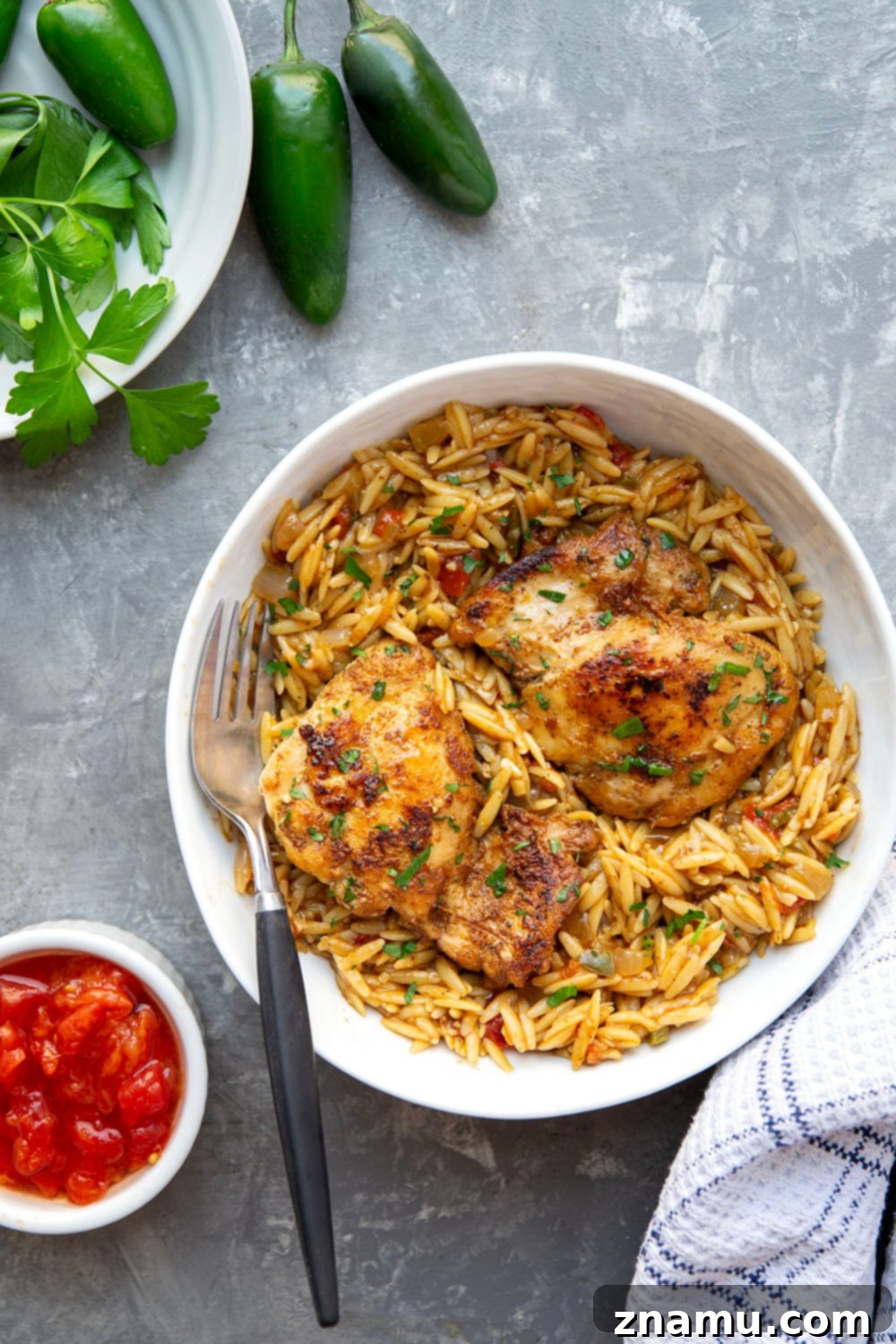 A close-up of a seared blackened chicken thigh, showing its dark, flavorful crust, placed atop a bed of creamy orzo. The background is slightly blurred.