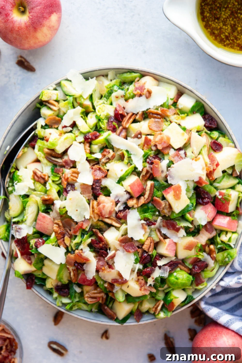Overhead shot of the ingredients for the warm Brussels sprouts salad arranged on a wooden board, including fresh Brussels sprouts, red apples, pecans, dried cranberries, bacon, and a bowl of maple mustard dressing.