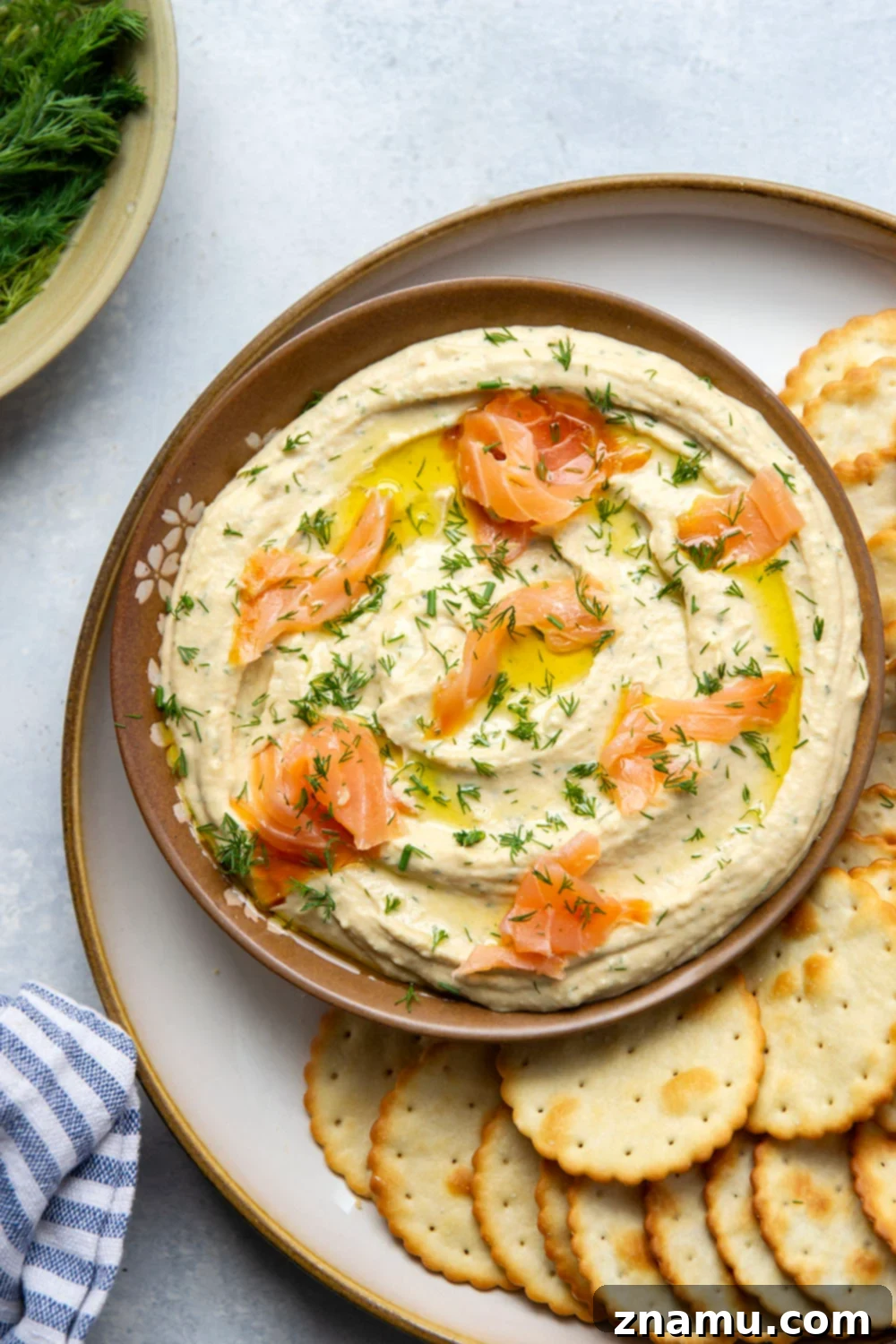 A hand dipping a toasted pita triangle into a bowl of Dill Smoked Salmon Hummus, with a blurred background of a relaxed setting.