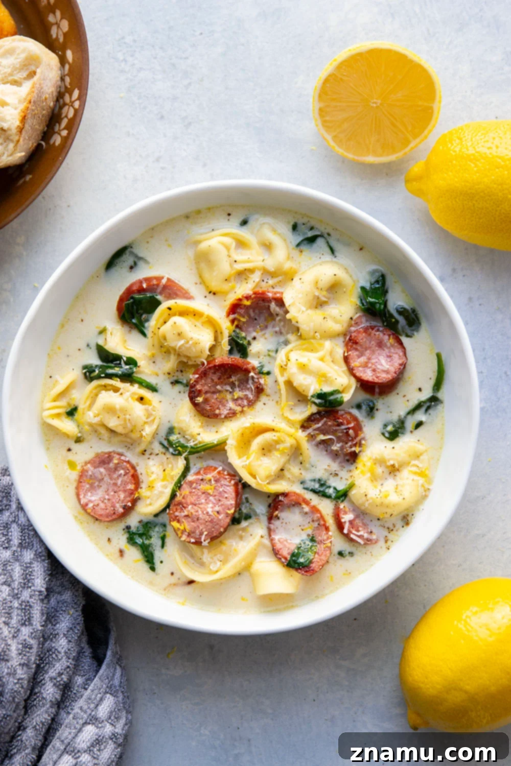 Sunshine Lemon Tortellini Soup 4 An array of fresh ingredients for lemon tortellini soup, including lemons, baby spinach, cheese tortellini, chicken sausage, onion, and garlic, arranged on a wooden board.
