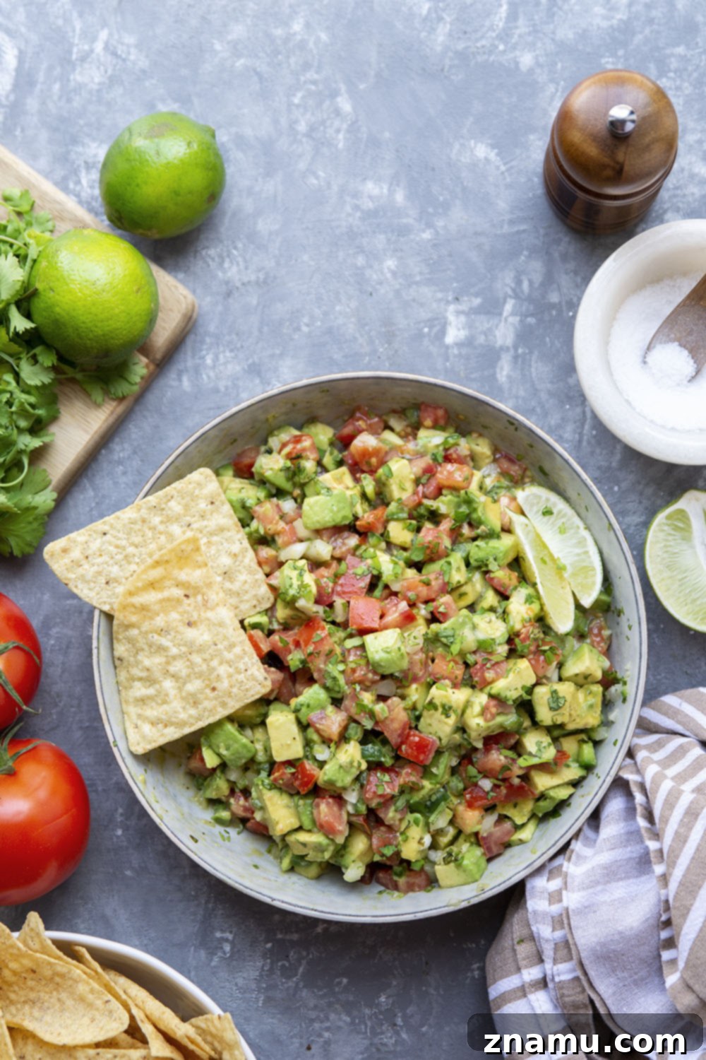The Ultimate Chunky Avocado Salsa 4 A bowl of fresh ingredients for chunky avocado salsa, including halved avocados, ripe red tomatoes, a lime, a jalapeño, and a bunch of cilantro, artfully arranged on a wooden board.
