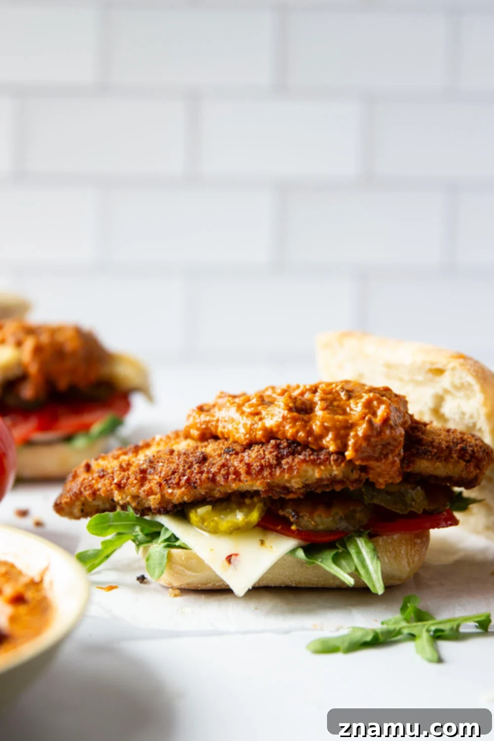 Romesco Chicken Crunch Sandwich 4 A close-up of a Crispy Chicken Romesco Sandwich, showcasing the golden fried chicken, vibrant red romesco sauce, and fresh green arugula inside a ciabatta bun.