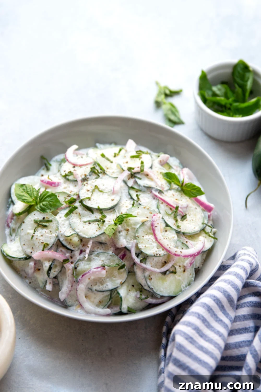 Velvety Basil Cucumber Delight 4 A close-up of a hand gently tossing thinly sliced cucumbers and red onion in a large bowl, preparing them for the creamy dressing.