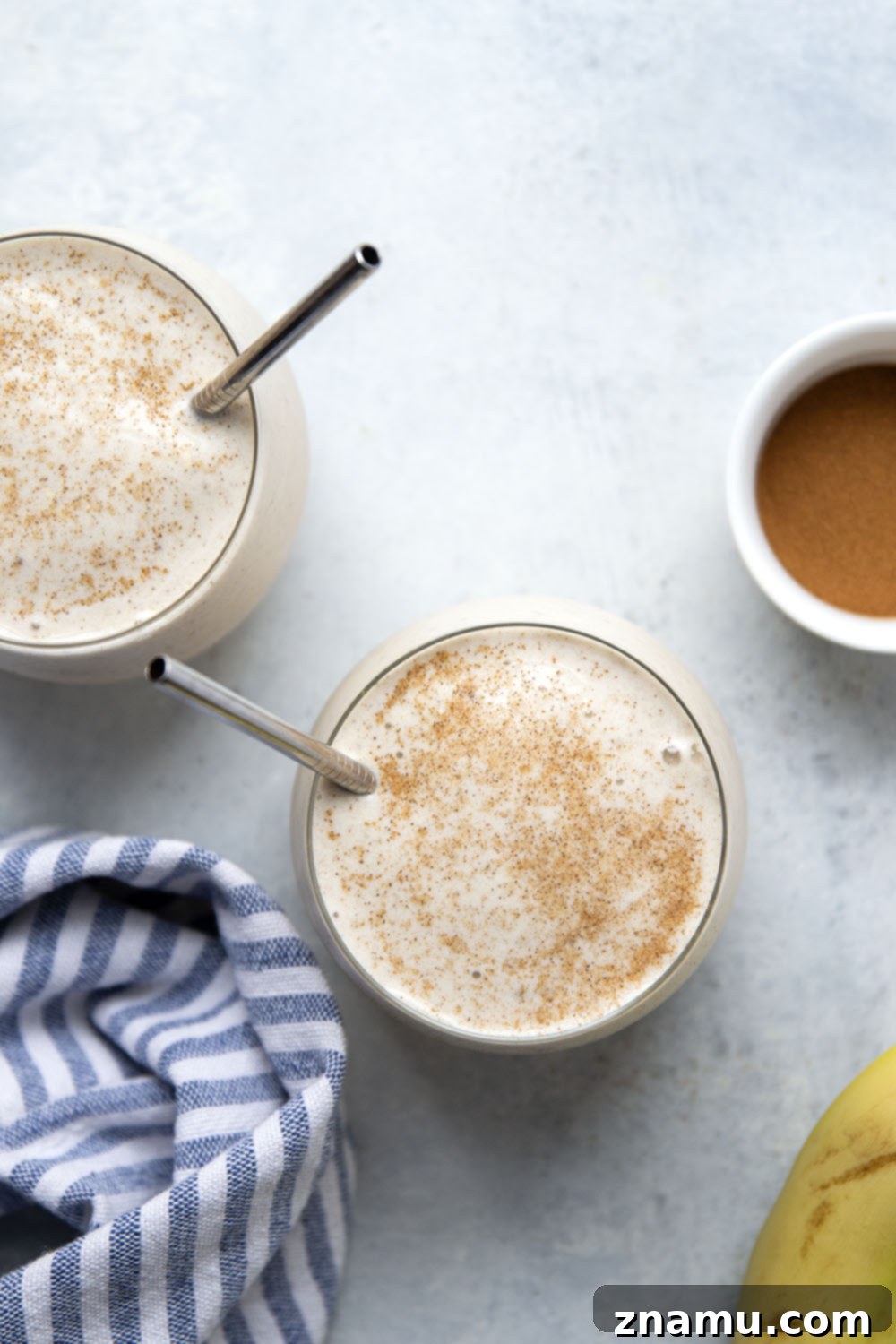 Spiced Banana Bliss Smoothie 6 A styled shot of a single glass of banana chai smoothie on a white surface, with a spoon and some scattered spices, emphasizing its appealing presentation.