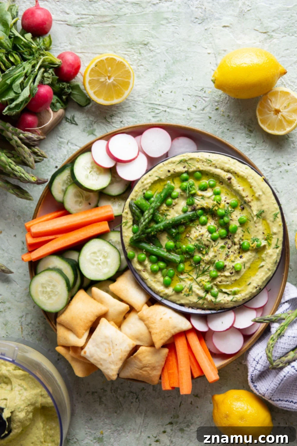 Emerald Asparagus Pea Hummus 5 A rustic wooden board with a bowl of asparagus pea hummus, surrounded by colorful fresh vegetables like cucumber, carrots, bell peppers, and pita bread for dipping.