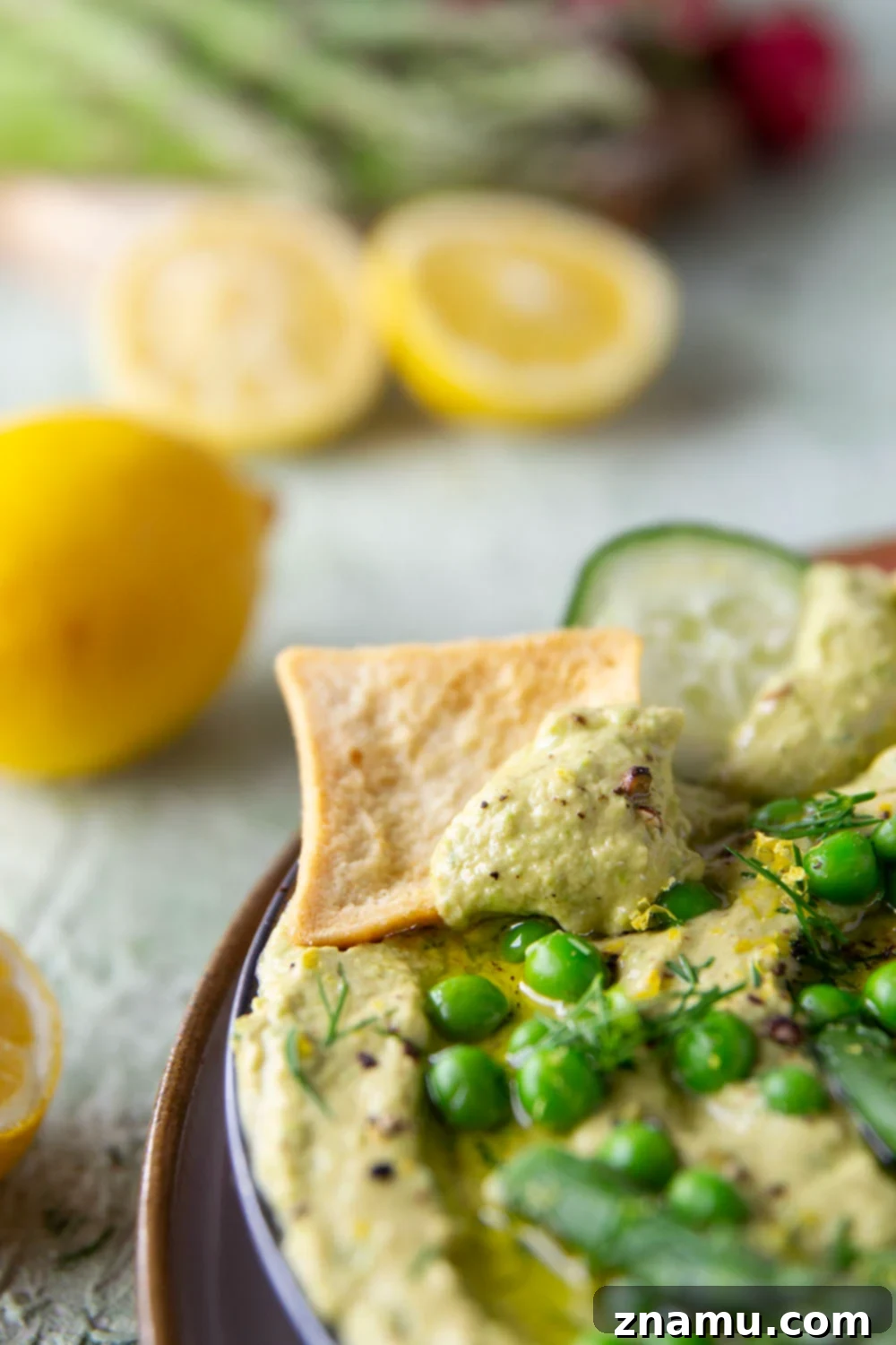 Emerald Asparagus Pea Hummus 6 A hand dipping a fresh cucumber slice into a bowl of creamy asparagus pea hummus, surrounded by other dippers.